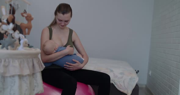 Happy Young Mother Lulls and Breastfeeds Her Little Baby on Fitball in Bedroom alt
