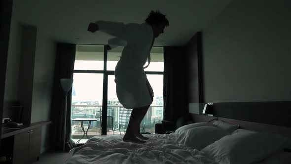 Happy Young Man Jumping on Bed at Hotel in Bathrobe alt