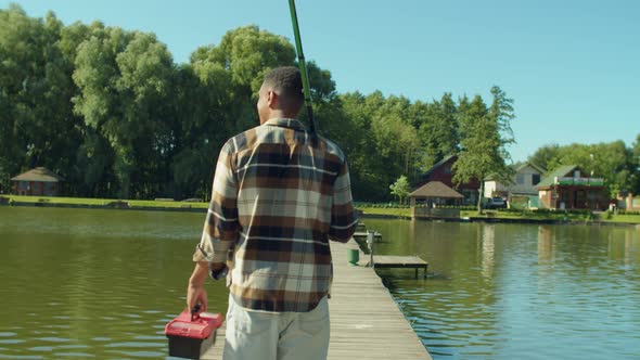 Rear View of African Fisherman with Rod and Tackle Box Walking on Pier on Lake alt
