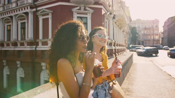 Two Young Smart Dressed Ladies in Sunglasses are Drinking Cold Tea Smiling Enjoying Summer Sunny Day alt