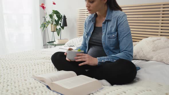 Caucasian woman in advanced pregnancy sitting on bed, eating salad and reading book. Shot with RED h alt