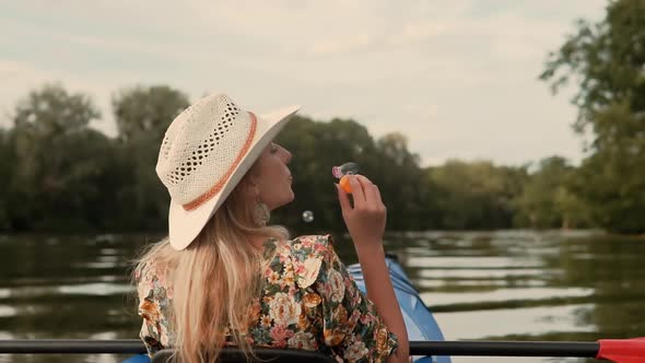Girl Enjoying At Sunset. Blonde Woman Blowing Soap Bubbles. Woman In Hat Relaxing On Kayak Boat. alt