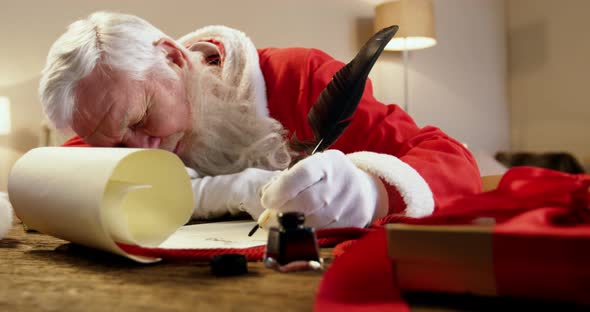 Santa Claus sleeping at desk while writing a letter with a quill alt