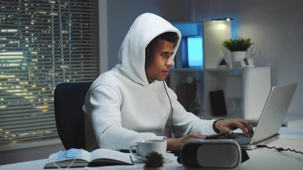 Gamer in White Hoodie and with Headphones Playing Games on Computer in the Evening alt