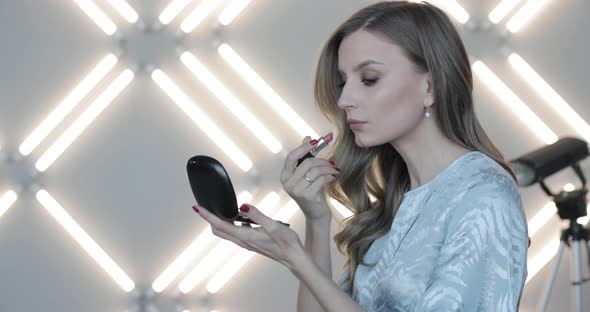 Beautiful Woman with Curled Hair Looking at Mirror and Painting Lips By Gloss