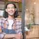 Male barista at cafe door, arms crossed, looks at the camera with welcome smile.