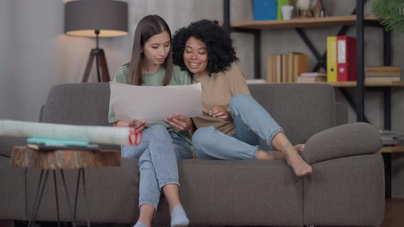 Young Cheerful African American Woman Entering Living Room with Focused Busy Asian Roommate alt