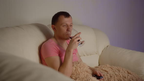 Young man in cosmetic mask drinking red wine and watching tv in the evening sitting on the couch alt