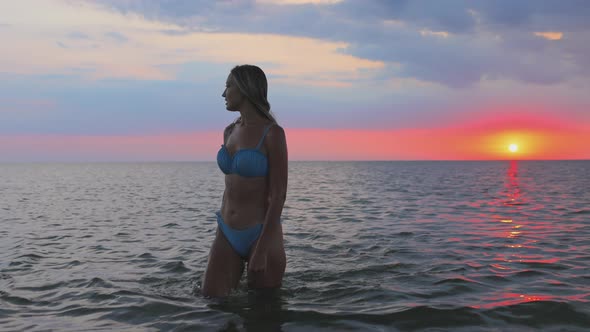 A Girl with Blond Hair is Going to Swim in the Lake at Sunset in the Evening alt