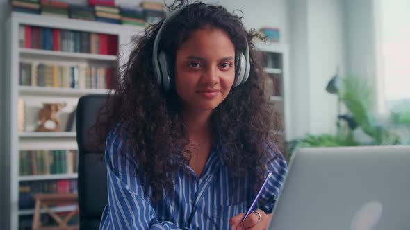 Young Indian Woman Student in Wireless Headphones Sits at Desk in Home Office alt