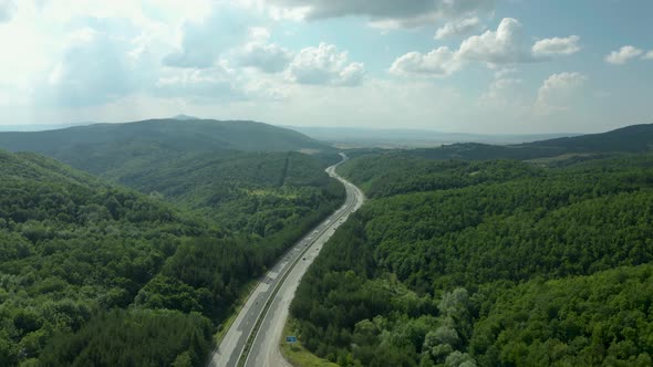 Highway road in green forest hills and blue cloudy sky, aerial pan up alt