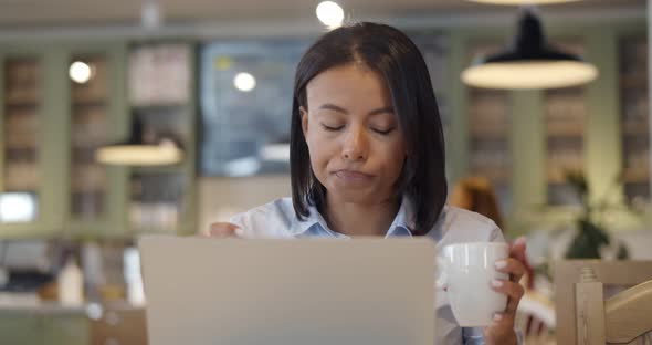 African Woman Sitting in Cafeteria Drinking Coffee and Working on Laptop alt