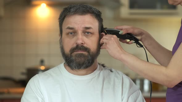 A woman cuts her husband's hair with an electric clipper in the kitchen during quarantine
