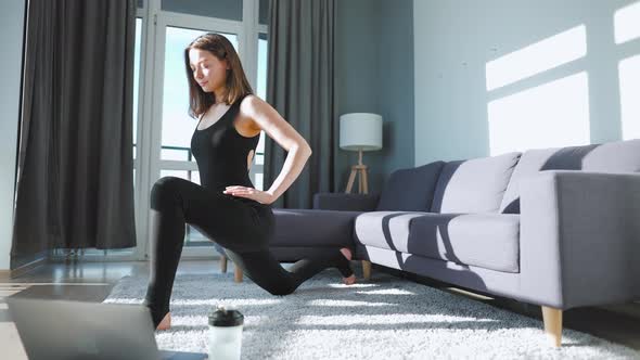 Young Caucasian Woman in Black Jumpsuit Doing Yoga at Home Online From Video Lessons on a Laptop alt