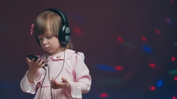 Little Girl in Big Headphones and Vintage Dress is Listening to Music on Phone alt