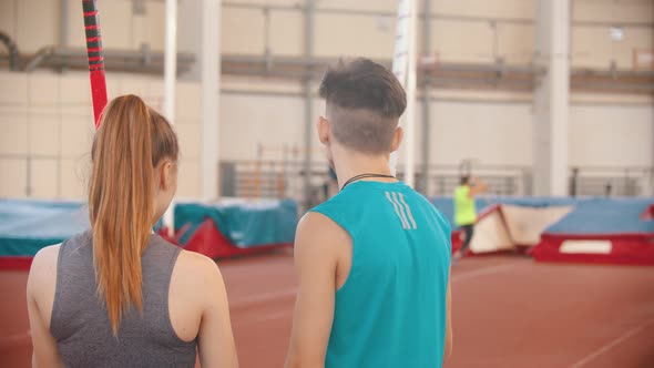 Pole Vault - Young Man and Woman Standing on the Stadium and Looking at Other Sportsmen Training alt