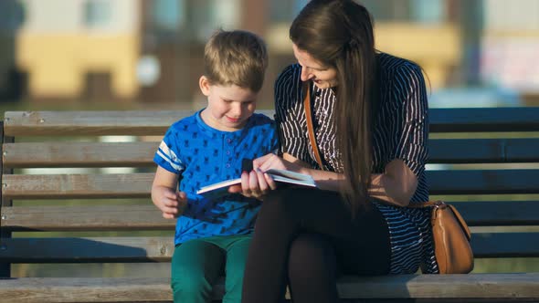 Young Mother and Son Sitting on a Bench in a Park and Reading a Book alt