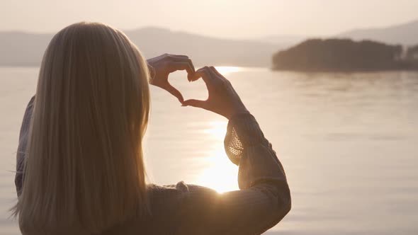 A Loving Blonde Woman Shows a Heart with Her Hands at Sunset By the Lake
