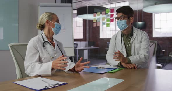 Diverse male and female doctors wearing face masks brainstorming in meeting room alt