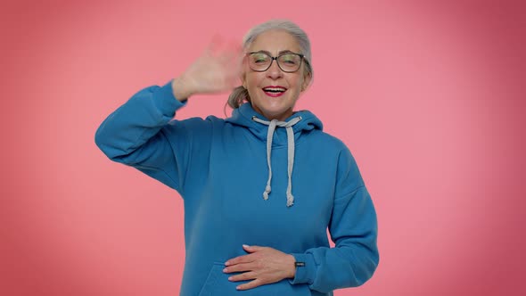 Happy Friendly Elderly Woman Waves Hand Palm in Hello Gesture Welcomes Someone Positive Emotions alt