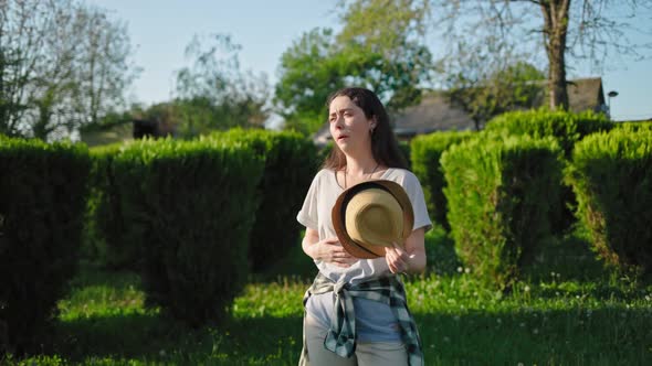 A young woman stands in a park fanning herself with a hat because of the heat alt