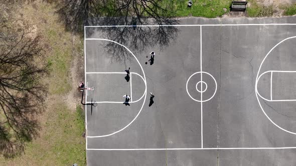 An aerial view over four people playing half court basketball surrounded by dry trees on a sunny day alt