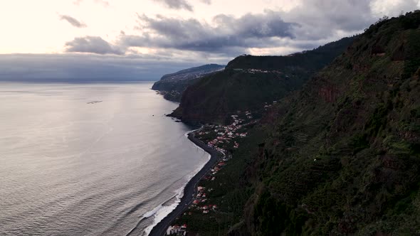 Sunset aerial along rugged Madeira coastline, small coastal village alt