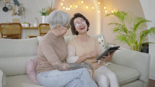 Two retired Asian senior female friends sitting on sofa using tablet together at home