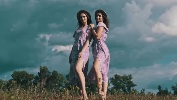 Two Young Slender Twin Girls in Identical Dresses Looking at Camera in Nature alt