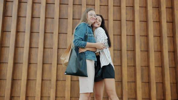 Happy Young Women Meeting Outdoors Hugging Talking Discussing Purchases in Shopping Bags alt