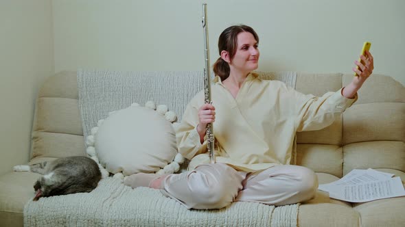 Happy woman musician with flute looking at mobile phone at home on sofa in living room