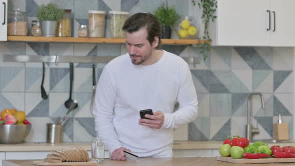 Young Man Making Online Payment on Smartphone in Kitchen alt