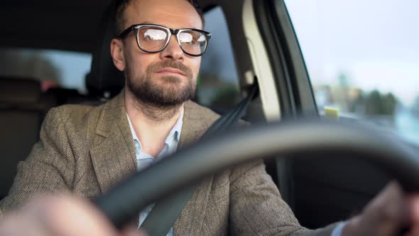 Satisfied Bearded Man in Glasses Driving a Car Down the Street in Sunny Weather alt