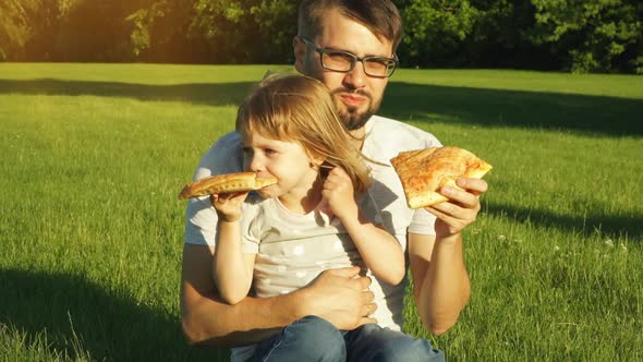 Father with Child Sit on Lawn in Summer Park at Sunset and Eat Pizza alt
