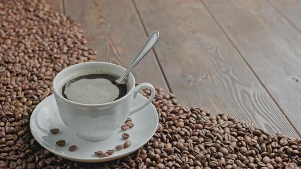 White Hand Stirring Black Coffee in a Mug on a Table Covered with Roasted Coffee Beans