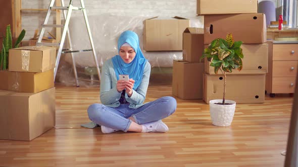 Young Muslim Woman in a Hijab Uses Smartphone Sitting on the Floor Next To the Boxes in a Modern alt