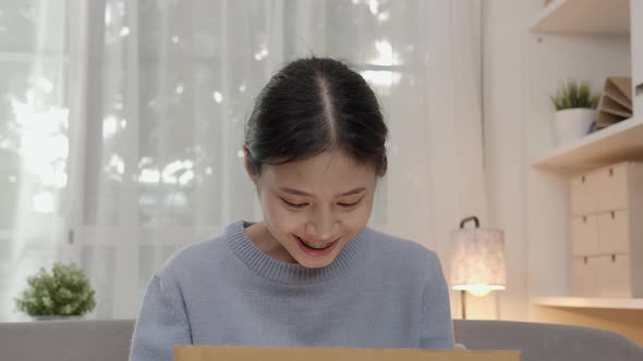 Young Asian woman open the cardboard box with surprise and sitting on sofa alt