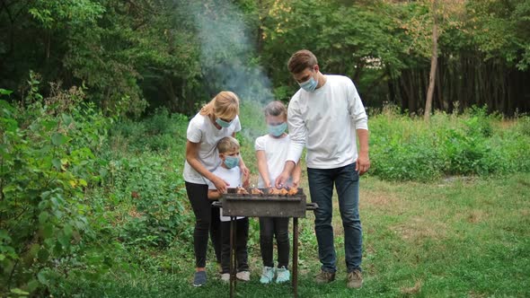 Dad, mom and children fry meat on skewers on the grill. Family in medical masks at a picnic