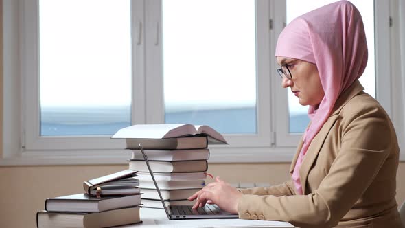 Side View of a Young Muslim Woman in a Pink Hijab Typing on a Laptop Among the Stacks of Books alt