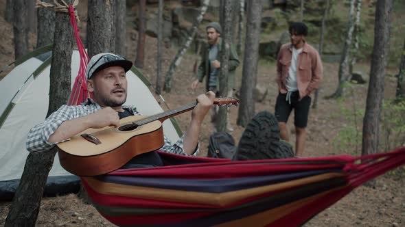 Cheerful Guy Playing Guitar Relaxing in Hammock in Campsite While Hikers Walking in Background alt