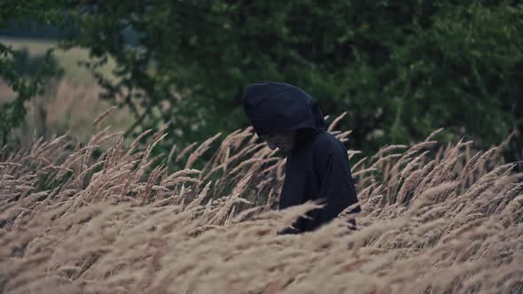 Scary figure with horrorful face outdoors. Ghost in black cloak walking in wheat field.  alt