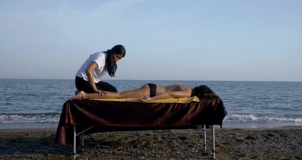 Female Master Is Massaging Naked Body of Woman Client Lying on Table on Sea Coast alt