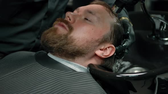 The Head of a Bearded Man is Washed with Shampoo Foam in a Barbershop alt
