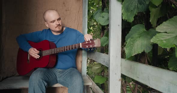Man with an Acoustic Guitar Sits on Porch of Farmhouse He Tunes the Instrument