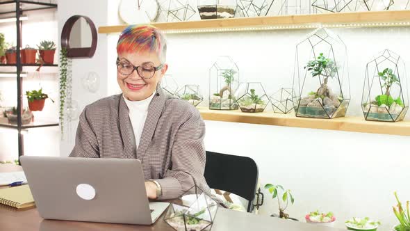 Mature Modern Business Lady Looking at Screen of Laptop Smile While Typing To Colleague alt