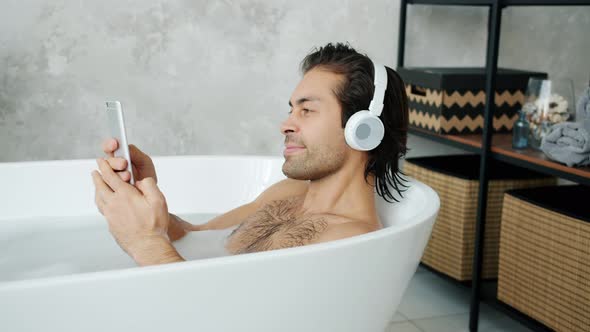 Guy Wearing Headphones Using Smartphone and Listening To Music in Bathtub at Home alt