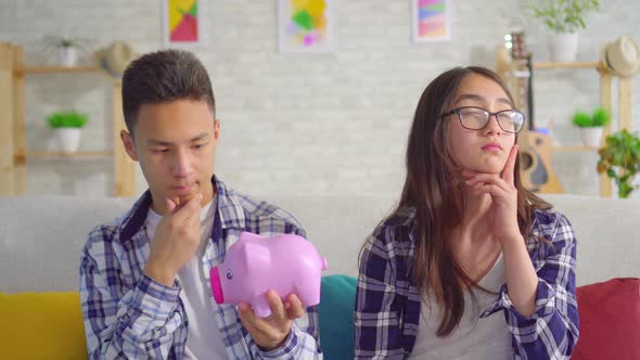 Thoughtful Young Sian Couple with Piggy Bank in Hand Sitting on Sofa alt