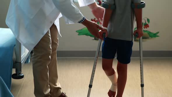Male doctor assisting injured boy to walk with crutches alt