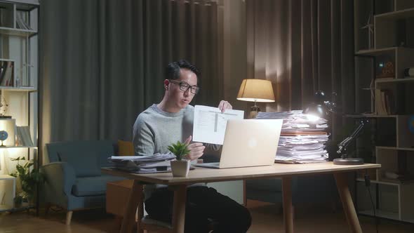 Asian Man With Documents Explaining Work On Paper While Having Video Call On Laptop At Home alt