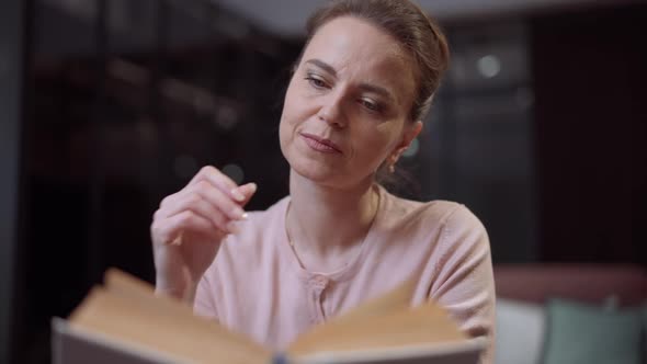 Focused Smiling Woman Turning Page Reading Book in the Evening at Home alt